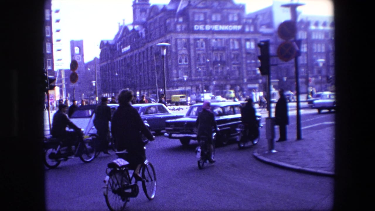 Amsterdam 1967 street traffic with several cyclists navigating among cars near traffic lights, with the De Bijenkorf building visible in the background.