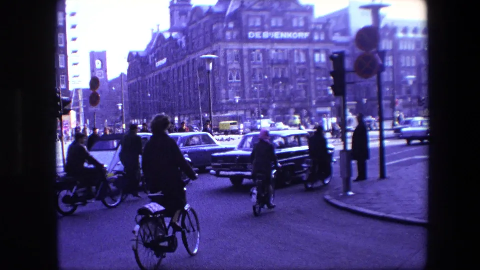Amsterdam 1967 street traffic with several cyclists navigating among cars near traffic lights, with the De Bijenkorf building visible in the background.
