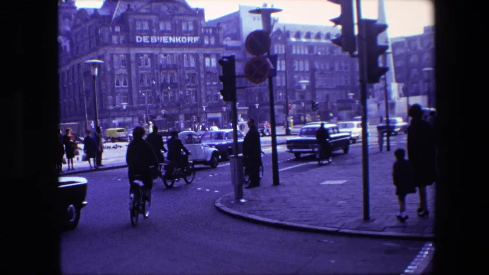 Amsterdam 1967 street scene with a cyclist riding toward traffic, pedestrians on the sidewalk, cars in the road, and the De Bijenkorf facade across the intersection.