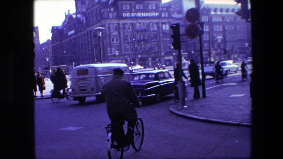 Amsterdam 1967 frame showing a cyclist from behind moving through mixed traffic with cars, a light van, pedestrians, traffic signals, and the De Bijenkorf building in view.