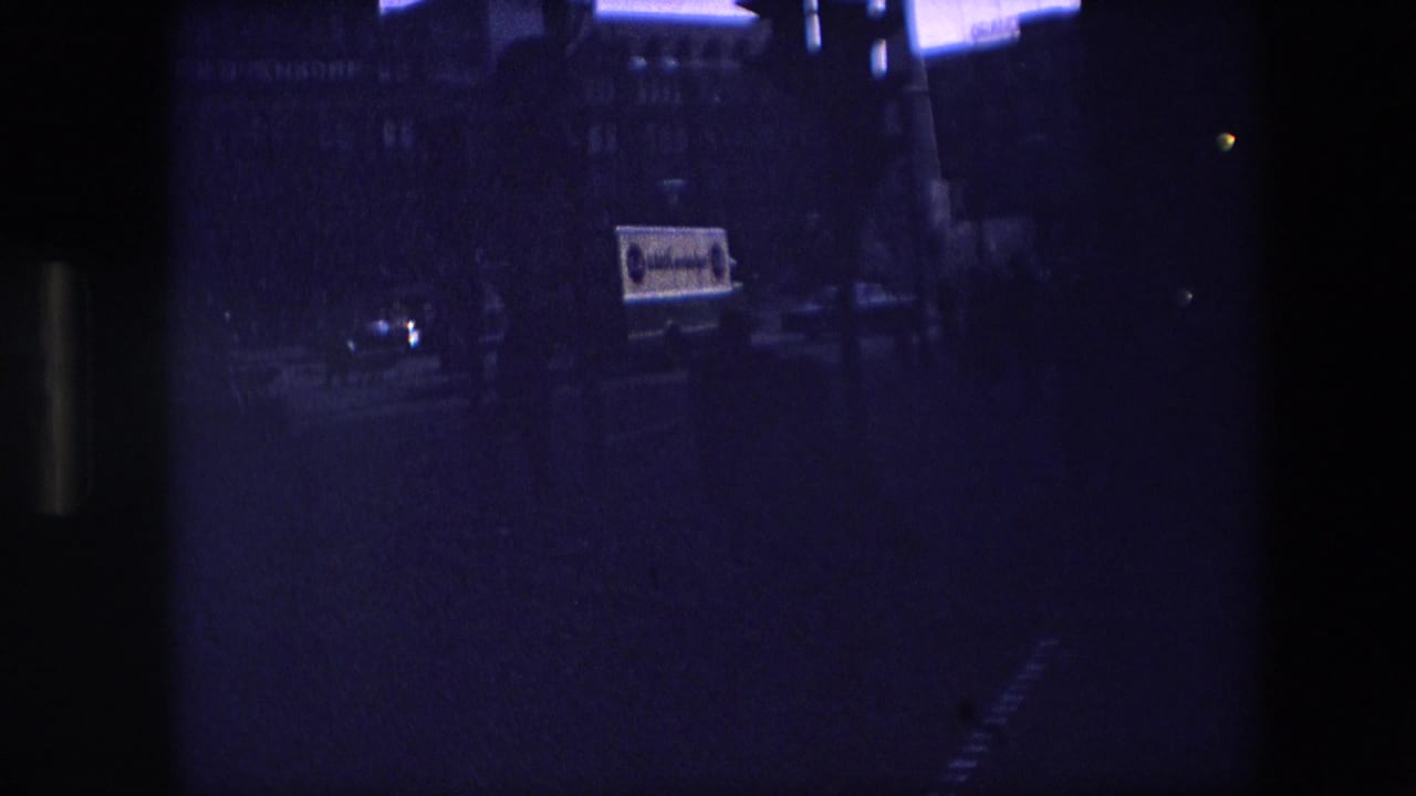 Underexposed 8mm frame from the clip, with a dim street scene, faint building outlines, and partial vehicle signage visible through heavy blue-purple shadow.