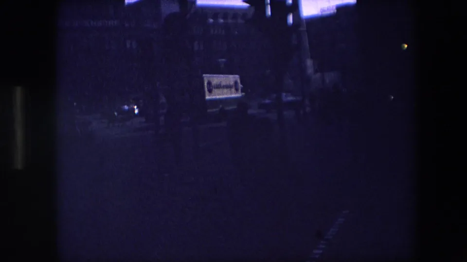 Underexposed 8mm frame from the clip, with a dim street scene, faint building outlines, and partial vehicle signage visible through heavy blue-purple shadow.
