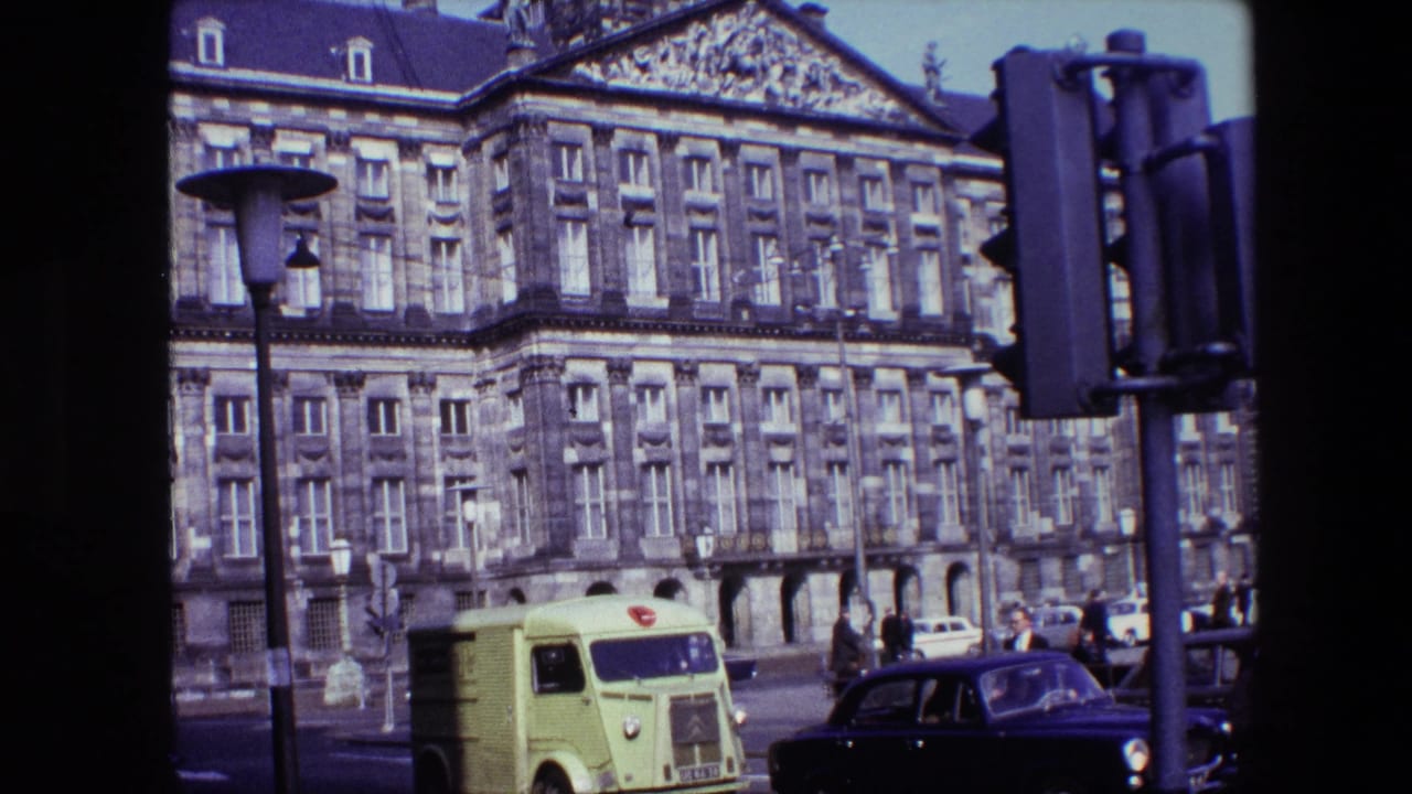 Ornate stone building behind a lime-green delivery van, dark car, pedestrians, lamppost, and traffic light.