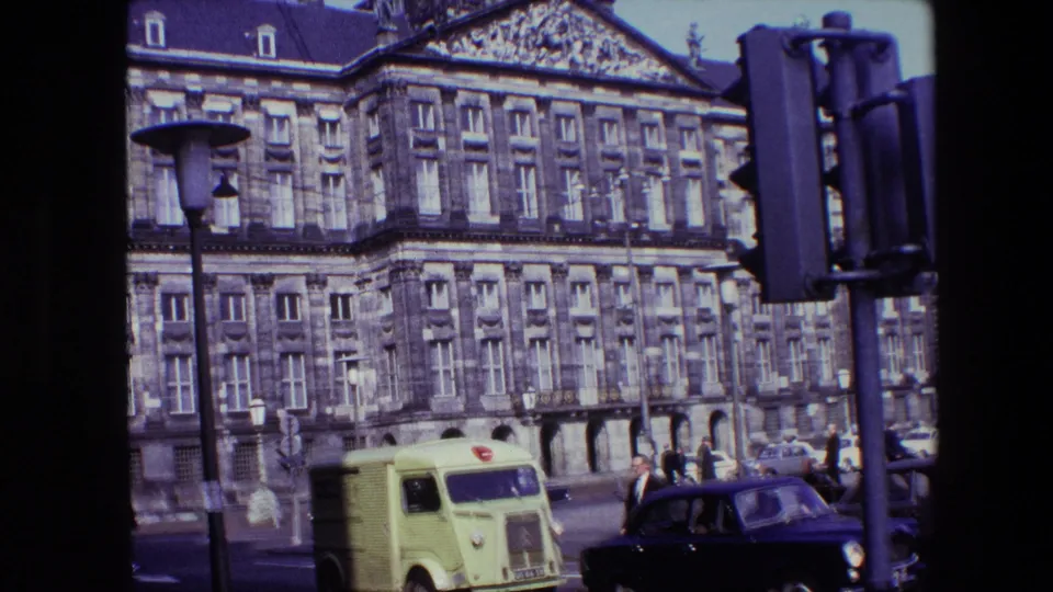 1967 Amsterdam street view with an ornate multi-story building, pole-mounted traffic lights, a lime-green delivery van, a dark classic car, and pedestrians moving near the curb.