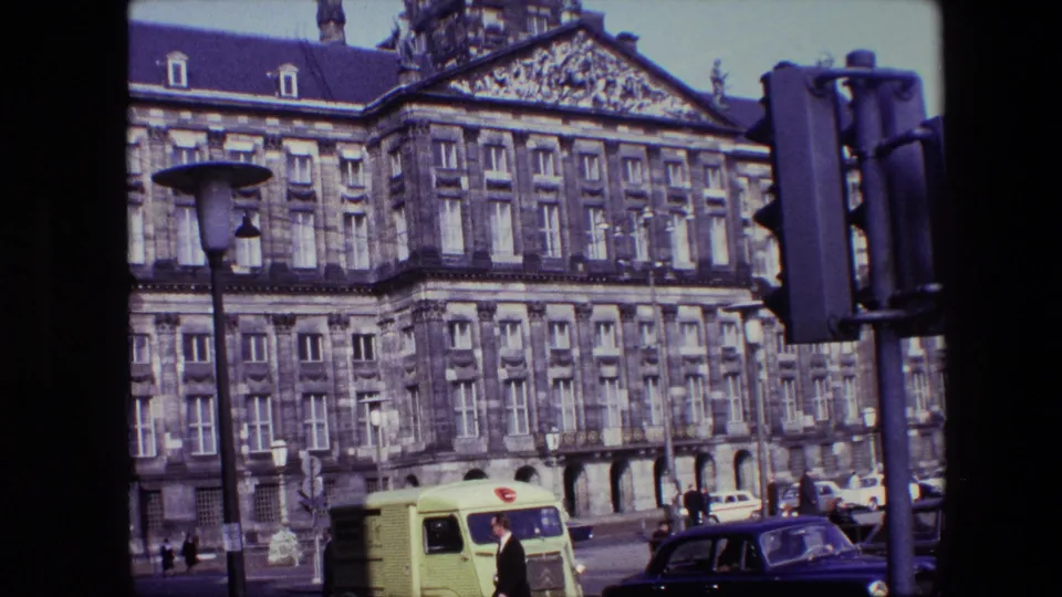 Final 8mm frame of the Amsterdam street scene, showing the ornate building facade behind a lime-green delivery van, dark blue car, pedestrians, lamppost, and traffic light.