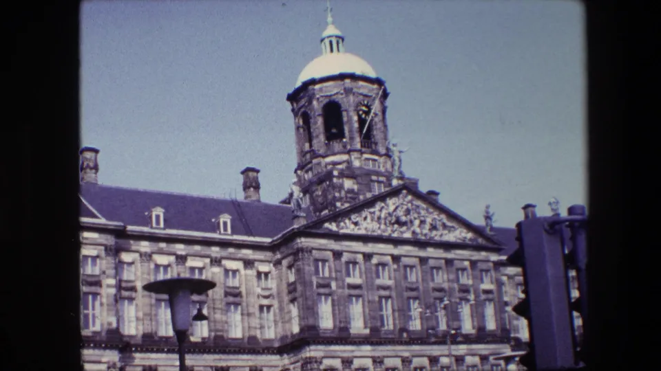 1967 8mm frame with the Royal Palace of Amsterdam tower rising above the roof, rows of windows below, and street fixtures crossing the foreground.
