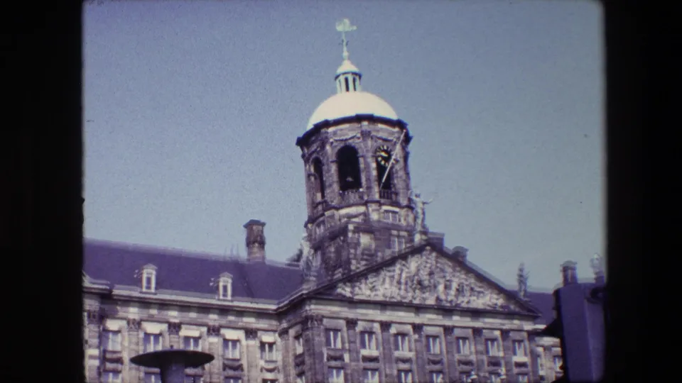 1967 8mm frame of the Royal Palace of Amsterdam upper facade and tower against a pale sky, with a street lamp and traffic signal at the edges.