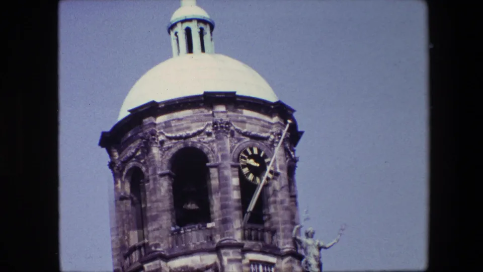 Tight 1967 Amsterdam home-movie frame showing the domed clock tower, clock face, arched opening, and upper stonework against the sky.