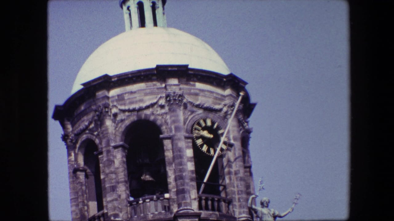 1967 8mm travel frame of an ornate Amsterdam clock tower with a pale dome, open bell arch, dark clock face with gold Roman numerals, and a diagonal flagpole.