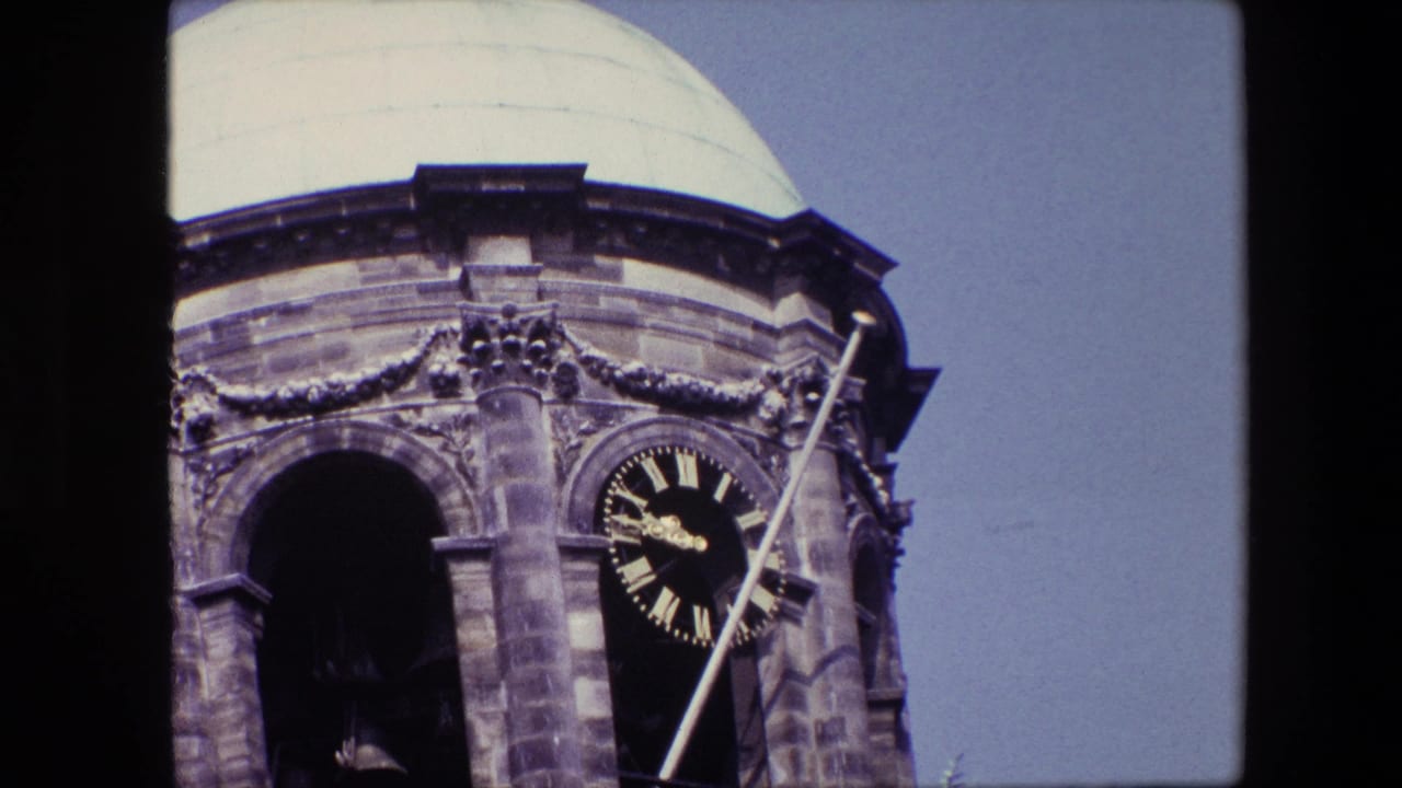 Closer 1967 8mm frame of the Amsterdam tower facade, showing carved stonework, an arched bell opening, a dark clock face, and a pale dome above.