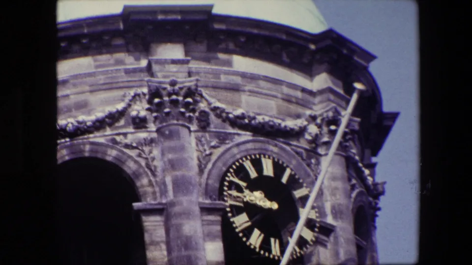 Zoomed 8mm frame emphasizing the tower's carved masonry, dark arched opening, gold Roman numeral clock face, diagonal flagpole, and pale dome edge.