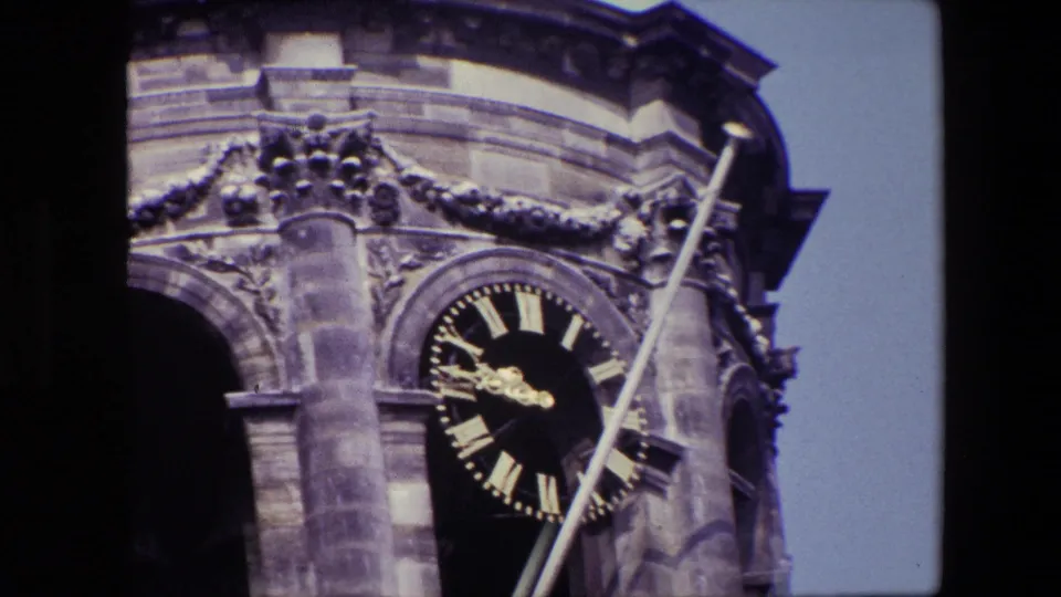 Tight 1967 Amsterdam travel frame showing the tower clock face with gold Roman numerals, surrounding carved stone, a dark bell arch, and the diagonal flagpole.