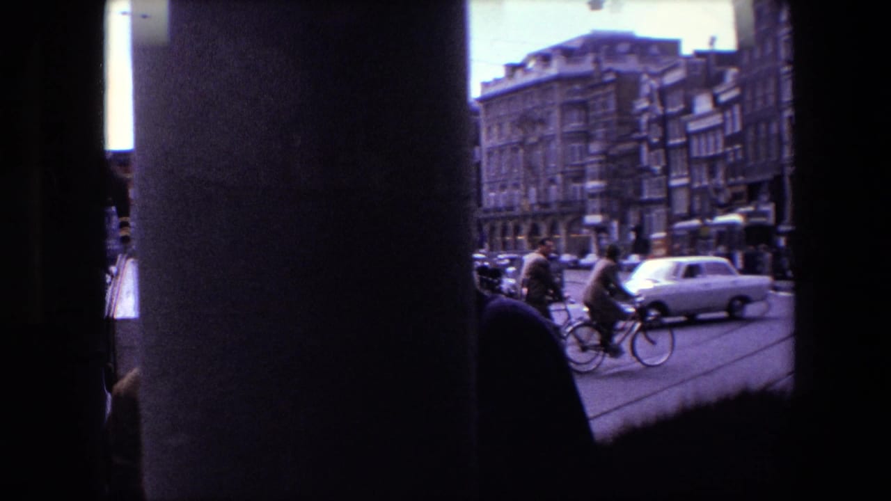 Amsterdam street traffic in 1967 seen through a dark obstruction, with cyclists, pedestrians, compact cars, and multi-story urban buildings visible.