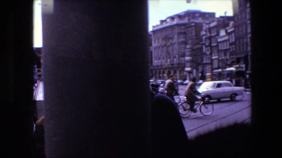 Amsterdam street traffic in 1967 seen through a dark obstruction, with cyclists, pedestrians, compact cars, and multi-story urban buildings visible.