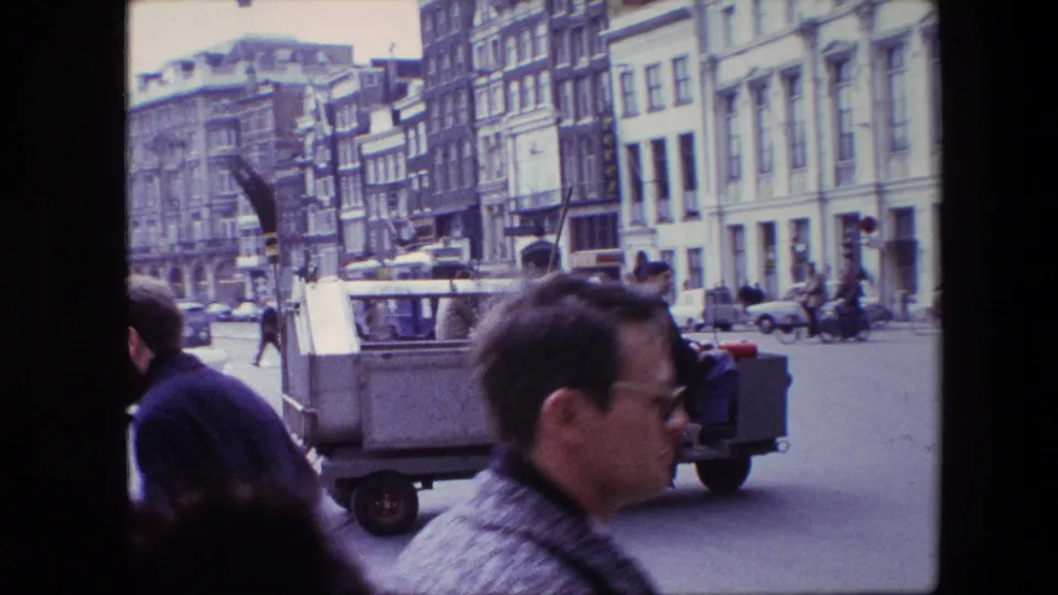 1967 Amsterdam street scene with pedestrians in the foreground, a small utility vehicle, parked or moving cars, and rows of older urban buildings.