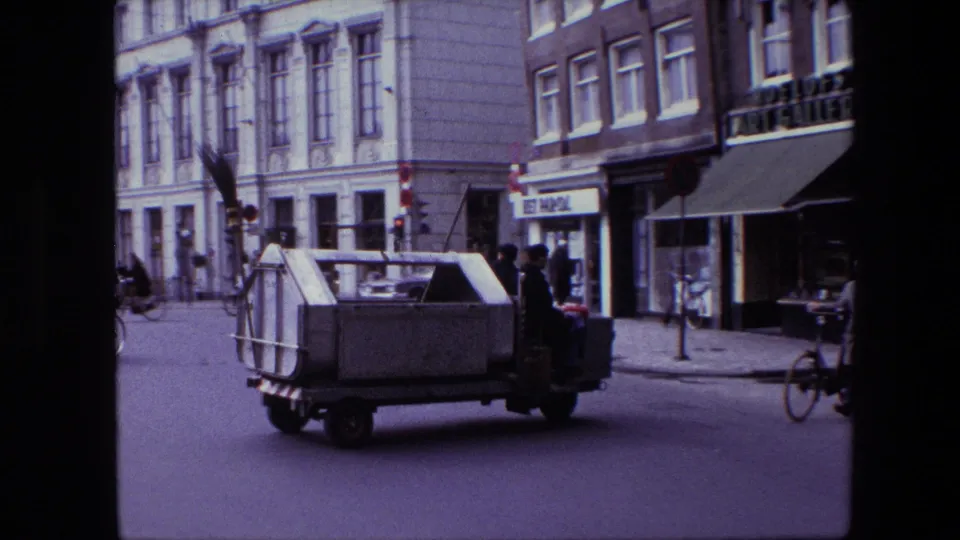 Small makeshift utility vehicle on a 1967 Amsterdam street, with a worker holding a broom, cyclists at the edge of frame, building facades, and shop signage nearby.