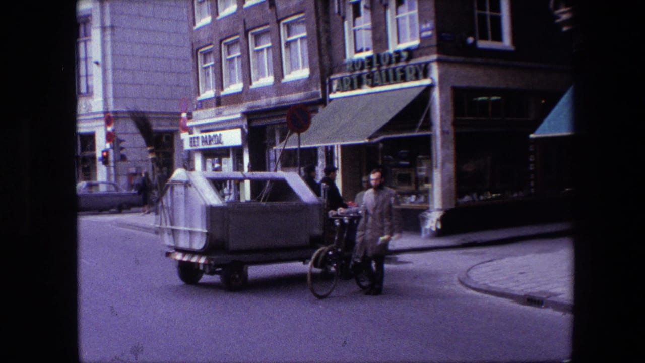 1967 Amsterdam street corner with a man beside a bicycle, a small utility vehicle, storefronts, and urban buildings visible in 8mm footage.
