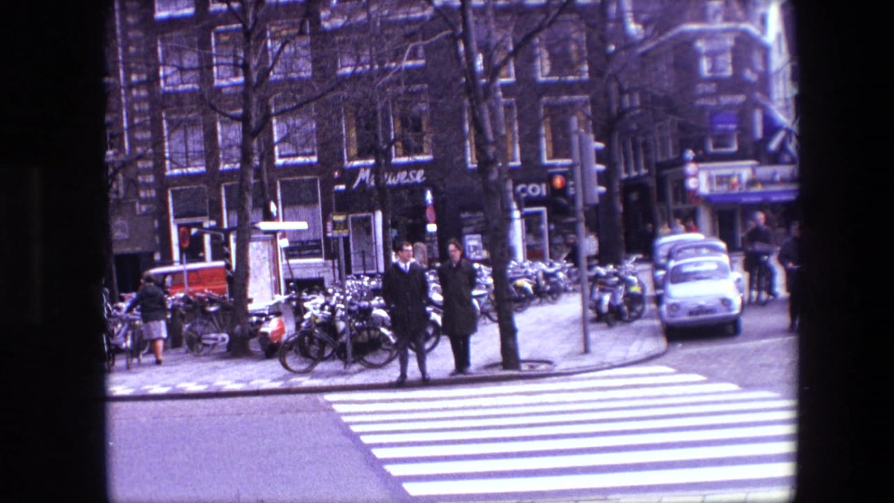 Pedestrians wait near a zebra crossing with parked bicycles and a small white car nearby.