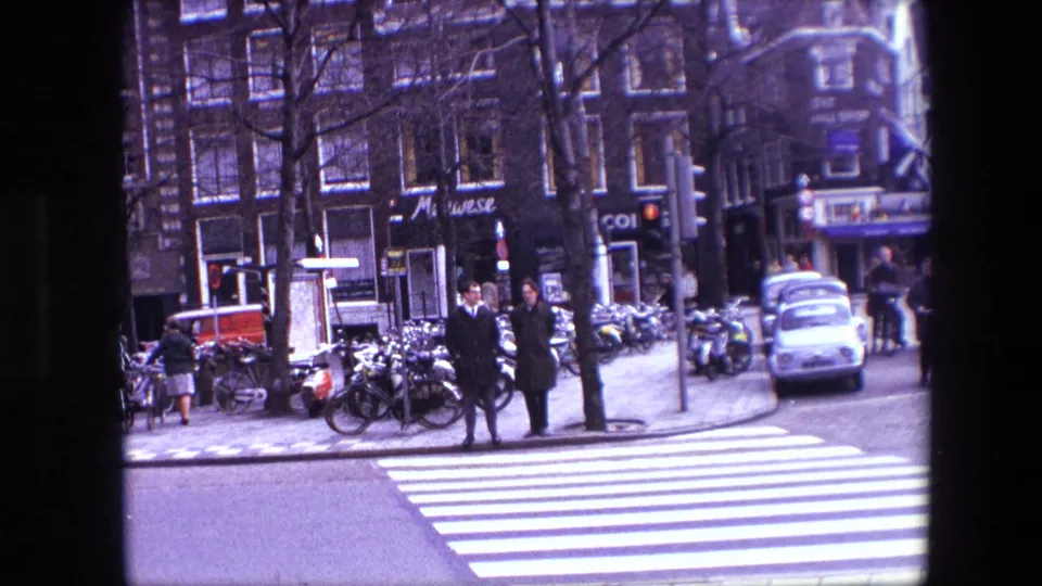 Amsterdam street view from 1967 showing pedestrians by a zebra crossing, rows of parked bicycles, storefront signage, and a small white car.