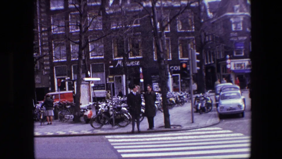 1967 Amsterdam street scene with pedestrians near a crosswalk, dense bicycle parking, storefronts, brick facades, and a compact car at the curb.