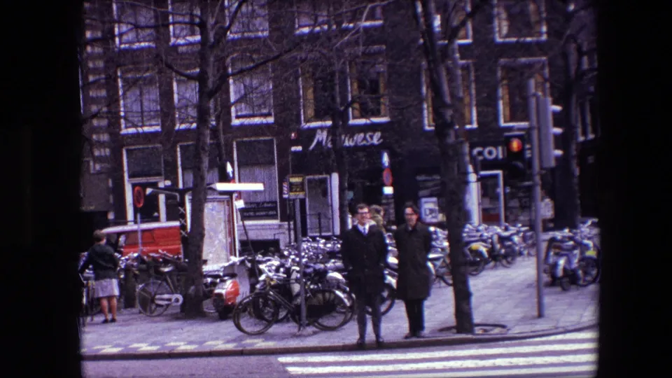 Amsterdam in 1967 with two pedestrians near a zebra crossing, bicycles along the sidewalk, storefront signage, and brick buildings in a home-movie frame.