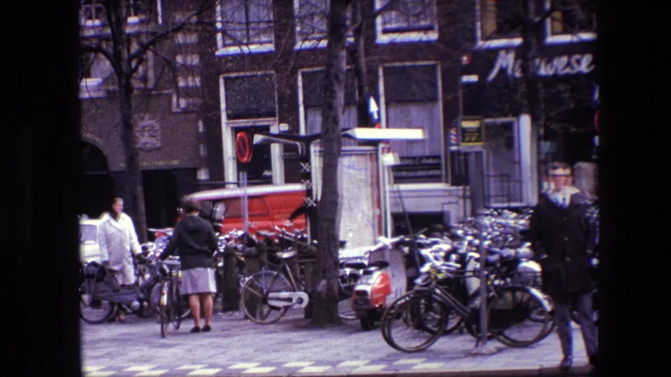 1967 Amsterdam sidewalk scene with people in coats, dense bicycle parking, a red van, storefront signage, and street furniture.