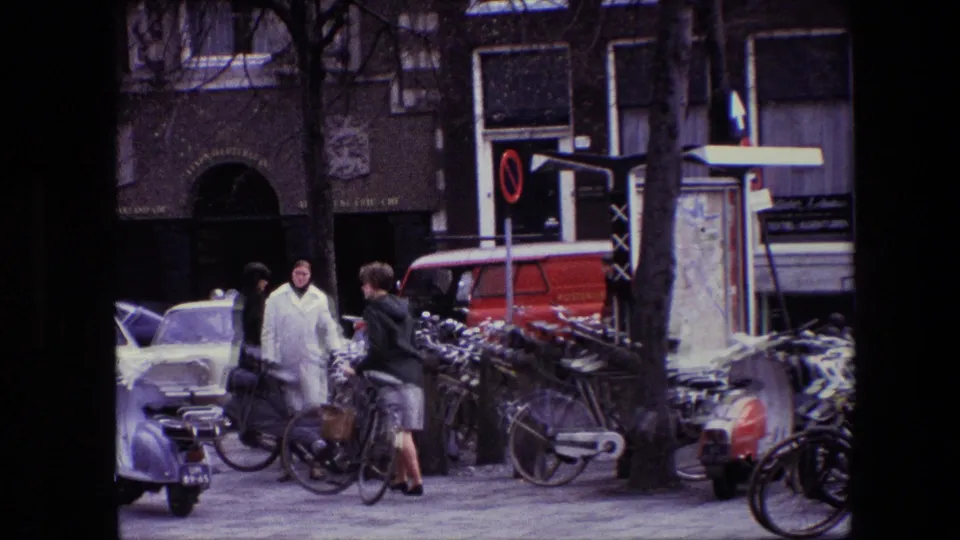 Amsterdam street frame from the supplied 1967 8mm clip showing people near bicycles, older cars, a red van, storefronts, and tree trunks.