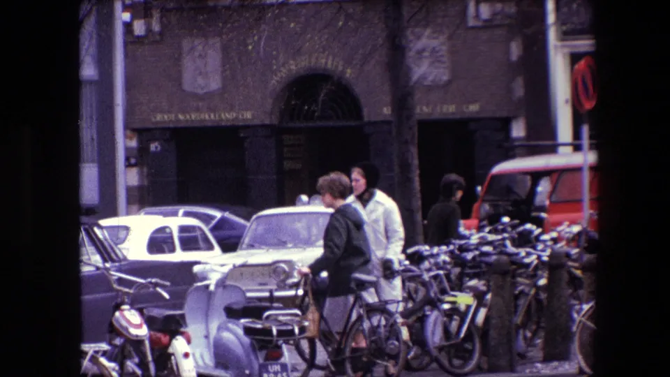 1967 Amsterdam street scene with pedestrians, bicycles, older cars, a red van, shopfront signage, and a handheld home-movie composition.