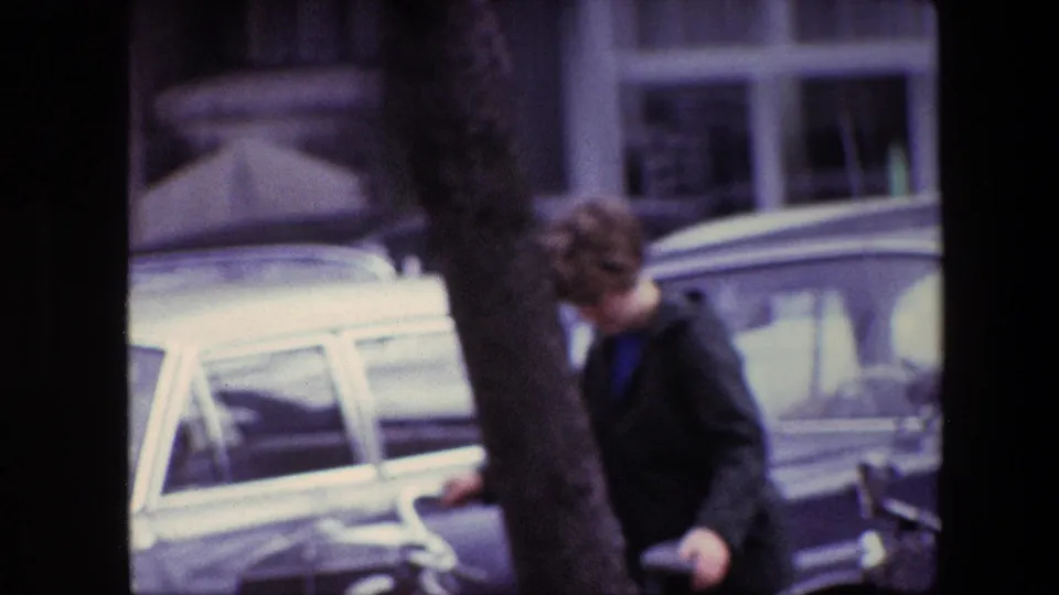 Soft 8mm frame from the supplied 1967 Amsterdam footage showing a person near a tree and parked cars at the edge of a city street.