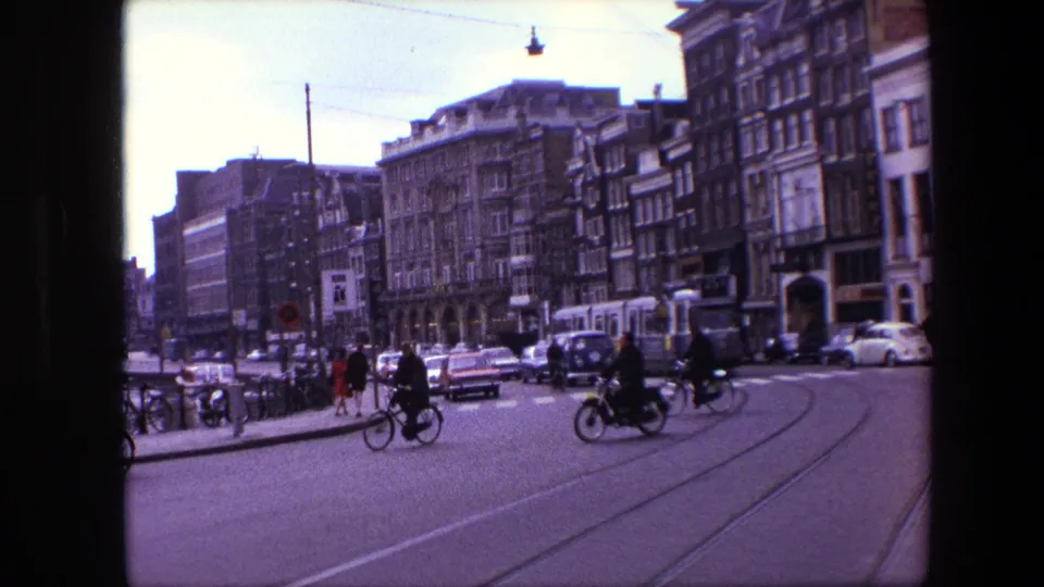 1960s Amsterdam intersection showing cyclists, pedestrians, stopped cars, tram rails, and historic streetfront buildings in a handheld 8mm frame.