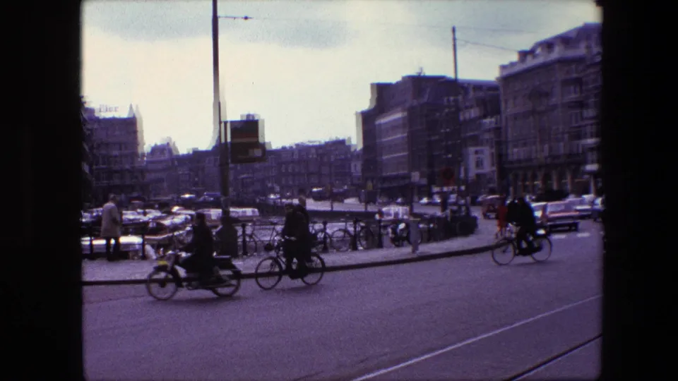 End frame of the supplied Amsterdam clip showing cyclists moving past parked bicycles, cars, tram rails, and distant city buildings under a pale sky.
