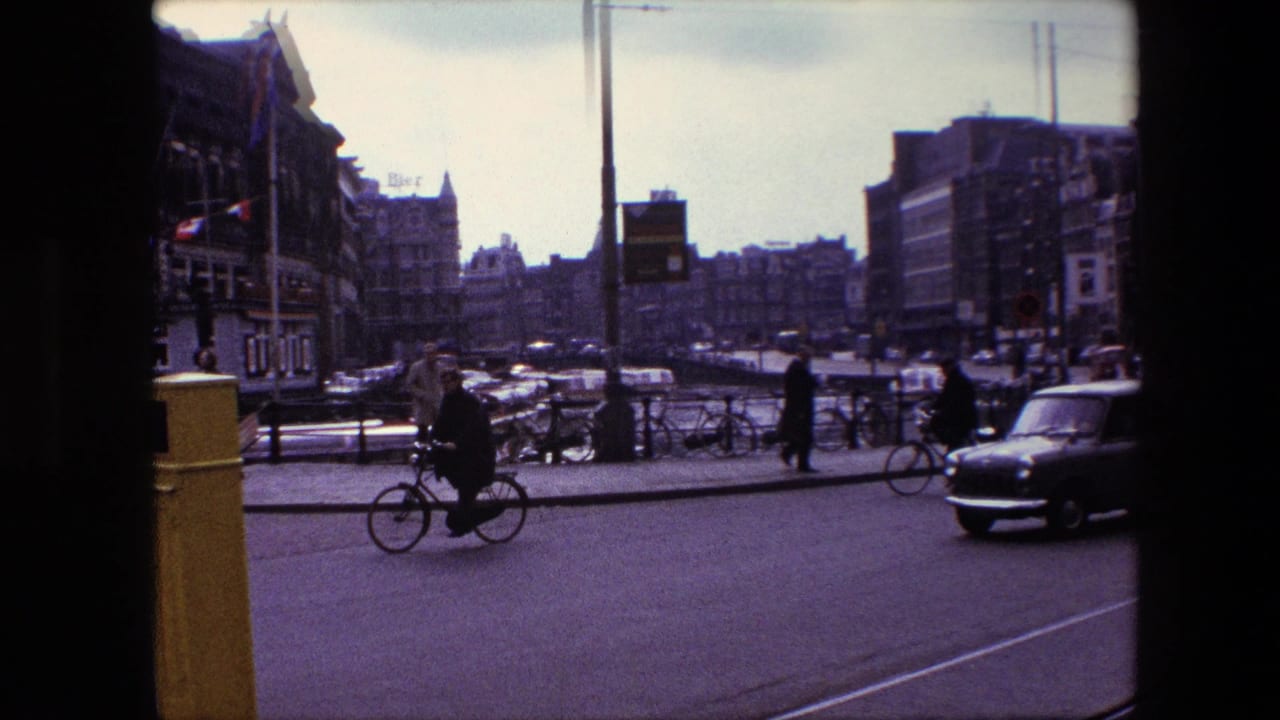 1967 Amsterdam street-corner view with a cyclist in the foreground, pedestrians near parked bicycles, cars moving through the road, and multi-story canal-side buildings beyond.