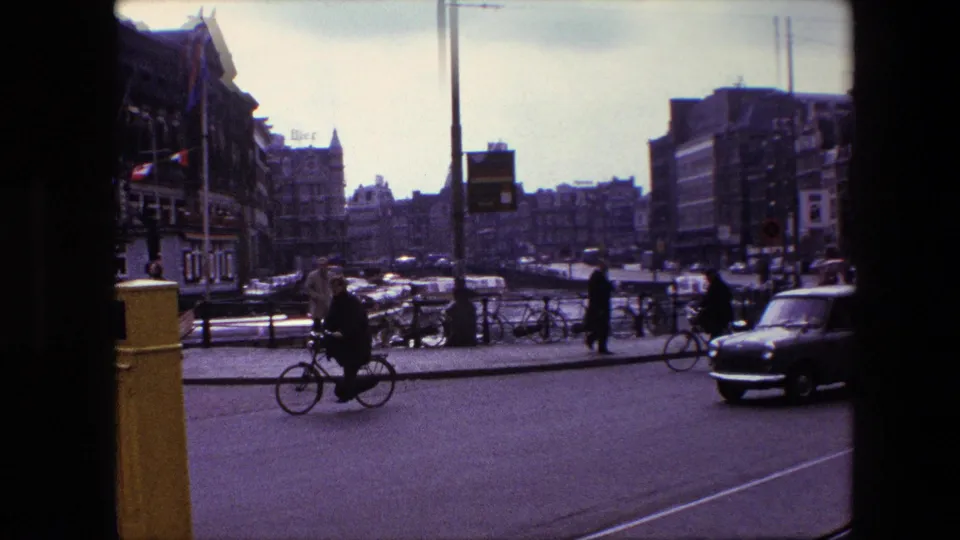 1967 Amsterdam street-corner view with a cyclist in the foreground, pedestrians near parked bicycles, cars moving through the road, and multi-story canal-side buildings beyond.