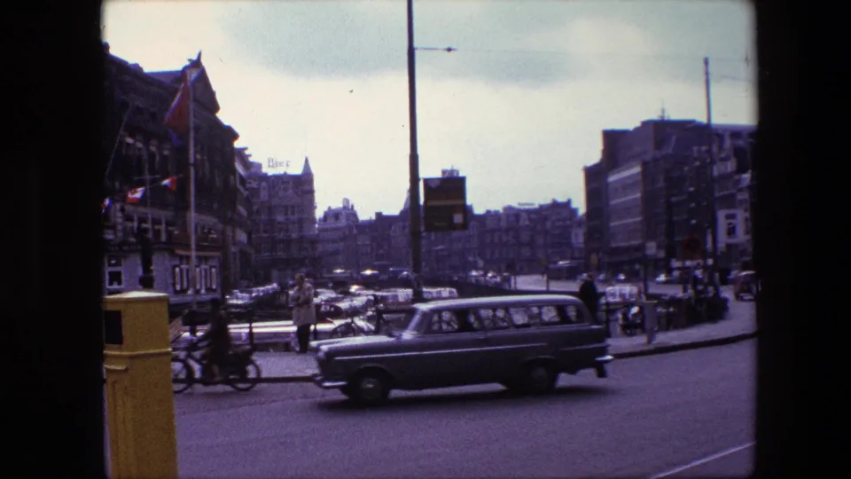 1967 Amsterdam home-movie frame showing a station wagon crossing the foreground while cyclists, pedestrians, parked bicycles, flags, and canal-side buildings fill the street corner.