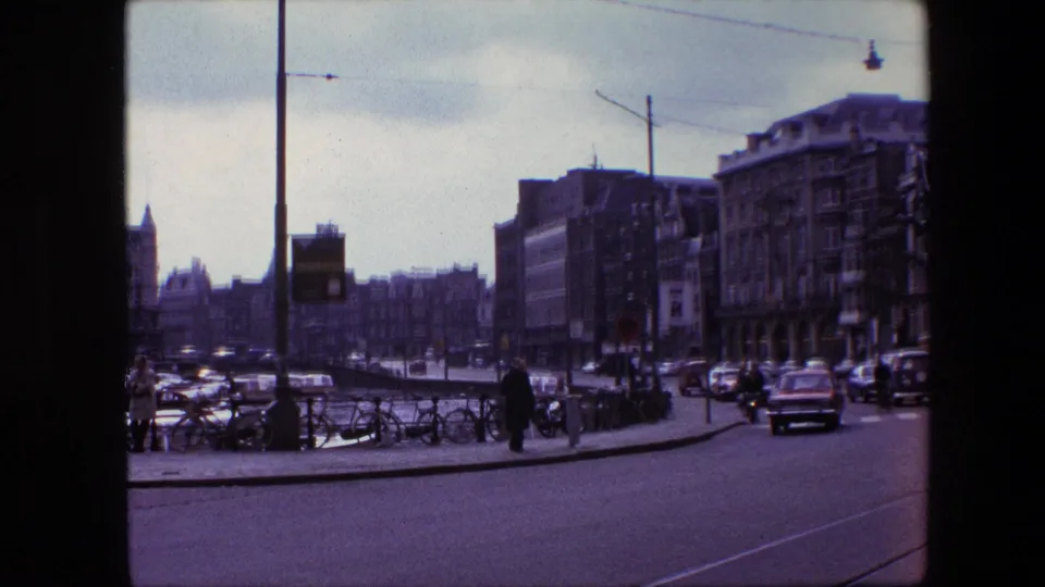 Wider 1967 Amsterdam street view with parked bicycles along a railing, pedestrians near the corner, cars on the road, tall street poles, and multi-story buildings under an overcast sky.