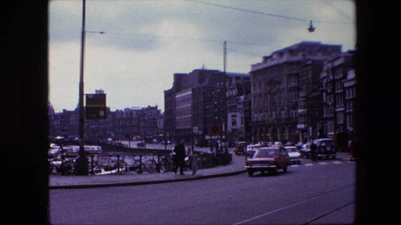 Amsterdam street traffic in the supplied 1967 context, with cars moving along tram tracks, bicycles parked by the curb, pedestrians on the sidewalk, and multi-story buildings beyond.