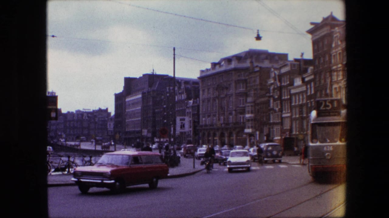 A red station wagon moves through a busy Amsterdam street scene, with tram rails, overhead wires, parked bicycles, pedestrians, and period buildings visible in the frame.