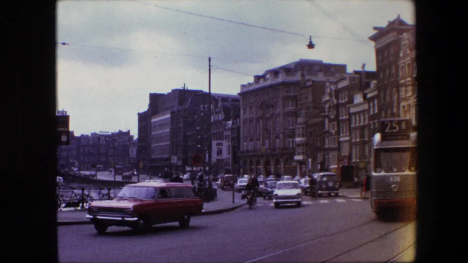 A red station wagon moves through a busy Amsterdam street scene, with tram rails, overhead wires, parked bicycles, pedestrians, and period buildings visible in the frame.