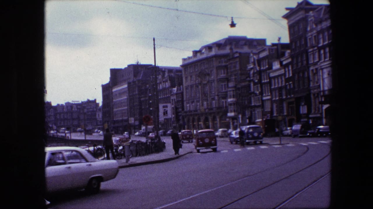 The frame shows 1960s Amsterdam street activity with cars entering the foreground, pedestrians near the curb, bicycles along the sidewalk, tram tracks, and older urban buildings.