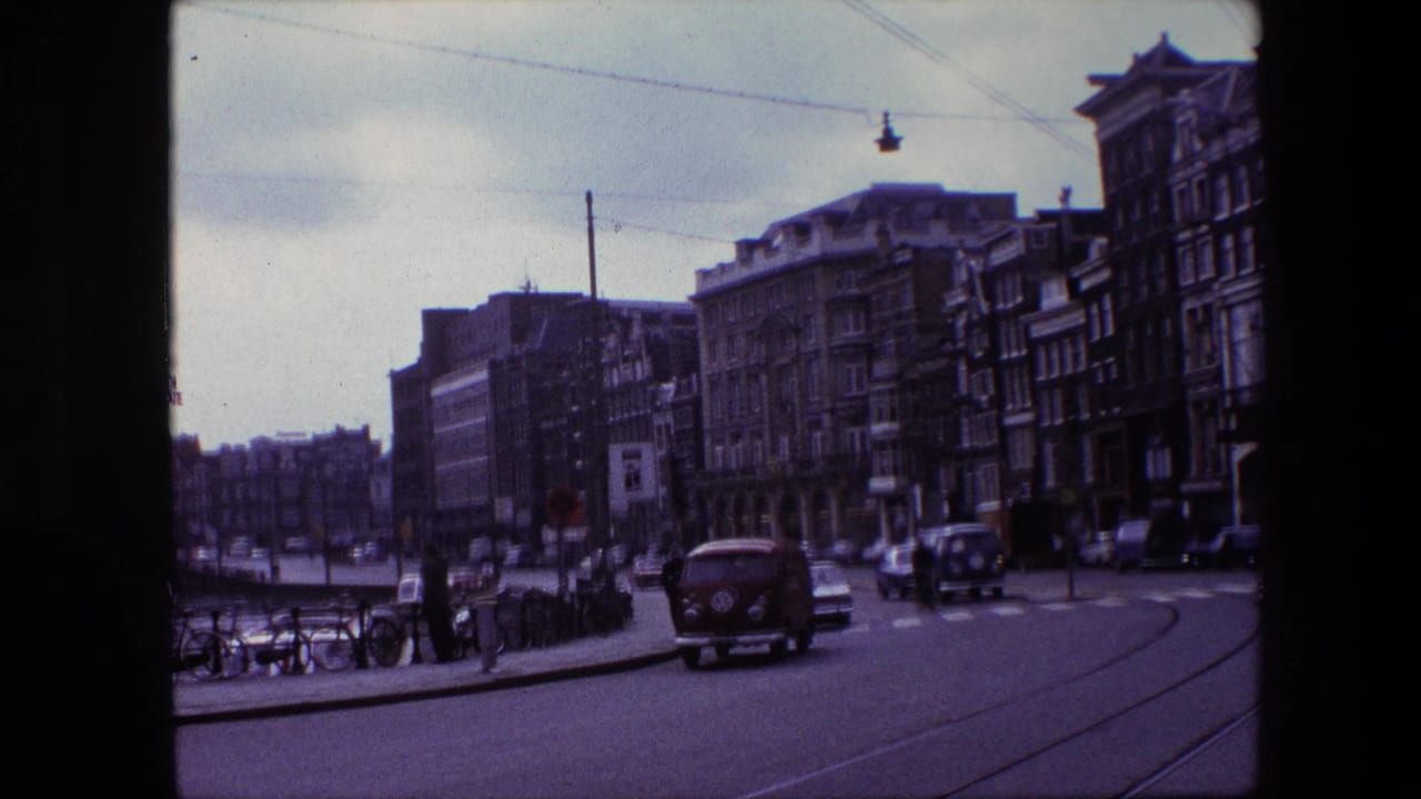 A red van travels away along tram rails in the Amsterdam street scene, with pedestrians, parked bicycles, cars, overhead wires, and multi-story buildings in view.