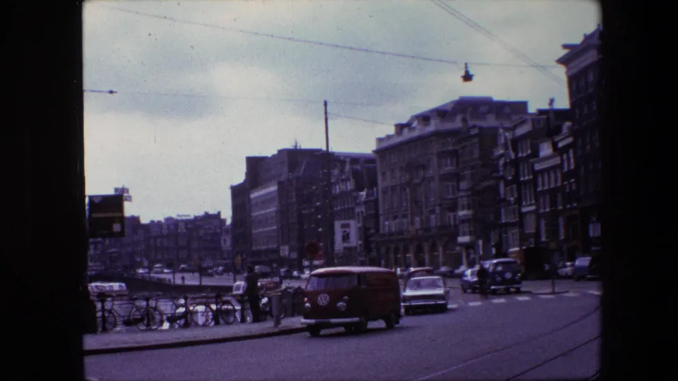 Amsterdam street scene from the supplied 1967 8mm clip, showing a red Volkswagen-style van, compact cars, parked bicycles, pedestrians, overhead wires, and multi-story buildings near a canal.