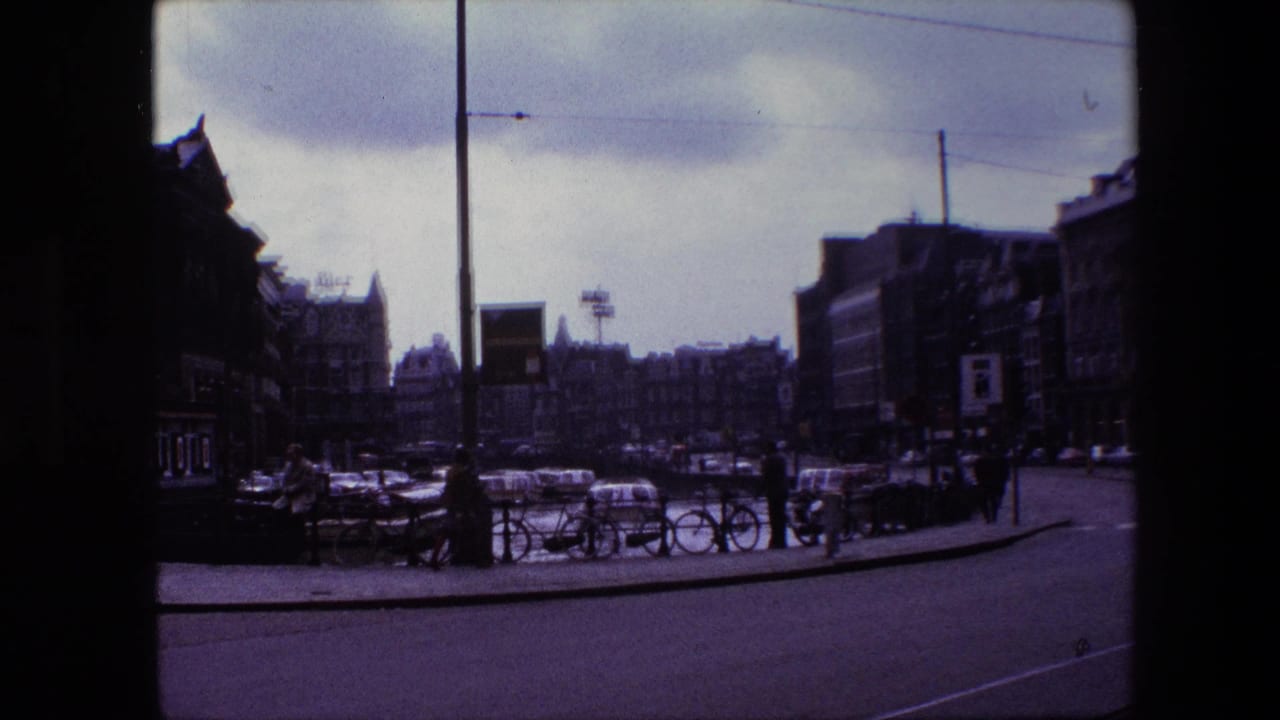 Darkened 8mm frame of an Amsterdam street corner from the supplied 1967 clip, showing parked bicycles beside canal railings, pedestrians, cars, overhead wires, and building facades.
