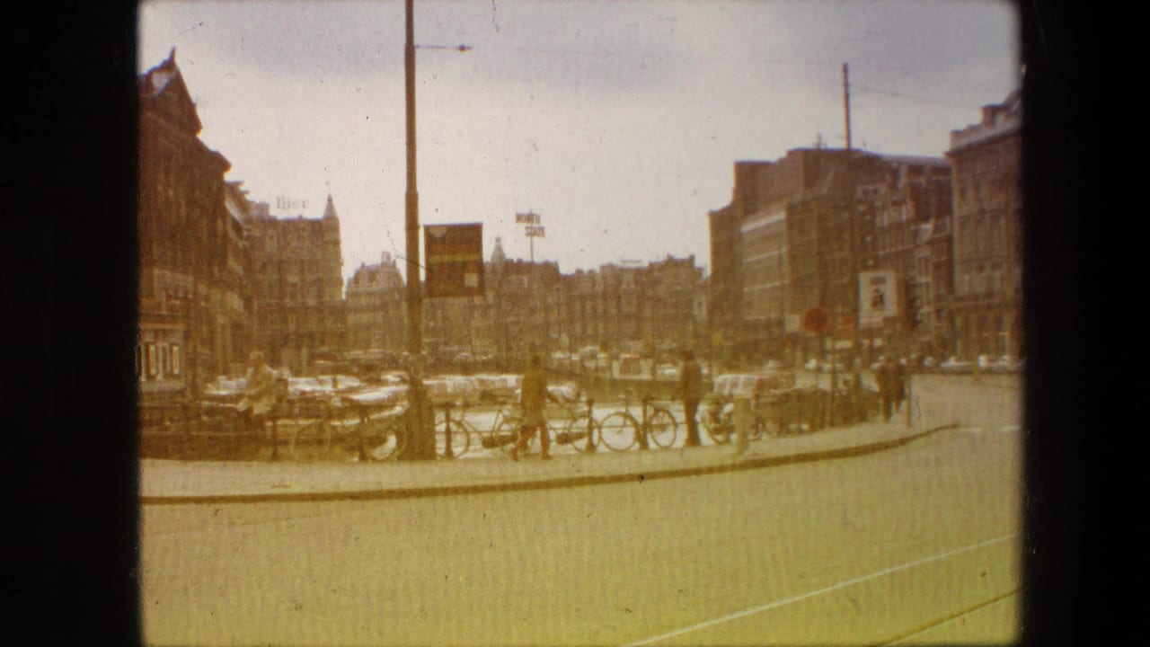 Faded 8mm view from the supplied 1967 Amsterdam footage, showing cars along a broad street, bicycles by canal railings, pedestrians on the pavement, overhead wires, and multi-story buildings.