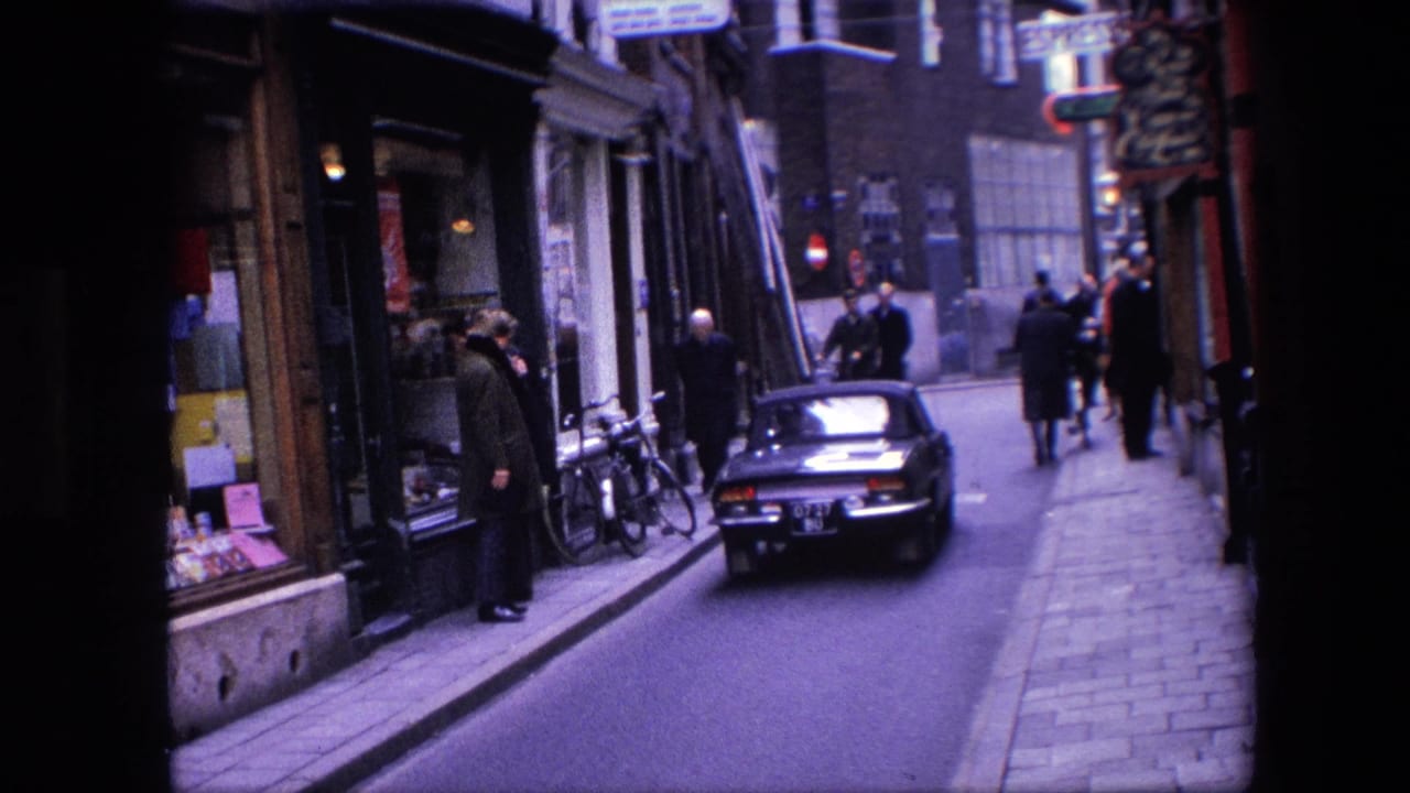 1967 Amsterdam street view with a classic car moving down the road, pedestrians near shop windows, bicycles at the curb, and period street signs overhead.
