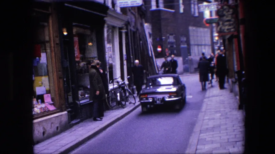 1967 Amsterdam street view with a classic car moving down the road, pedestrians near shop windows, bicycles at the curb, and period street signs overhead.