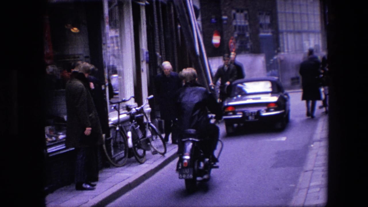 1967 Amsterdam street scene showing a motor scooter behind a car, pedestrians on the sidewalk, parked bicycles, shopfronts, and brick buildings.