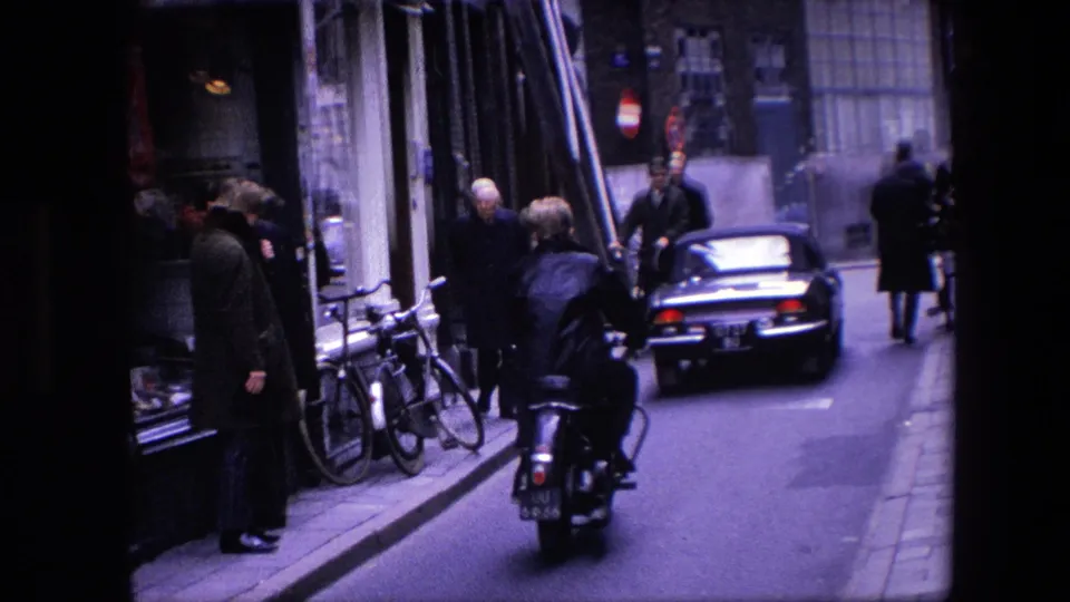 1967 Amsterdam street scene showing a motor scooter behind a car, pedestrians on the sidewalk, parked bicycles, shopfronts, and brick buildings.