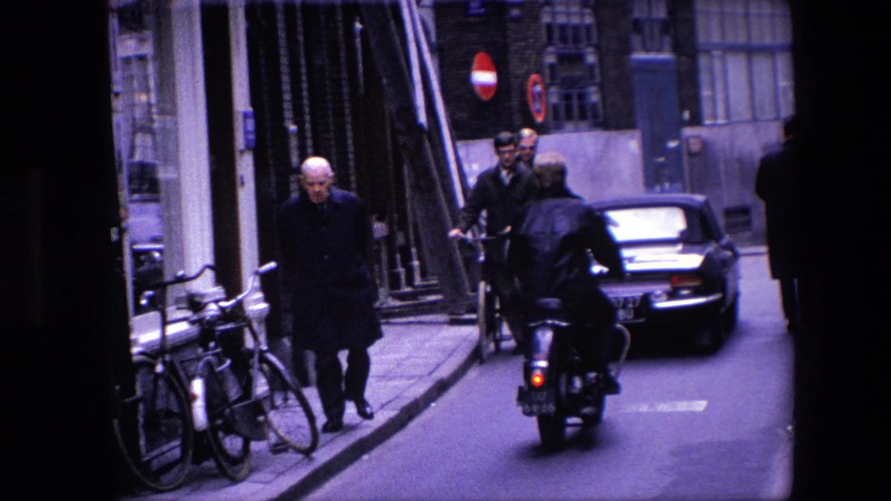 1967 Amsterdam street view showing a motorcycle rider following a dark classic car while pedestrians pass buildings and bicycles line the curb.