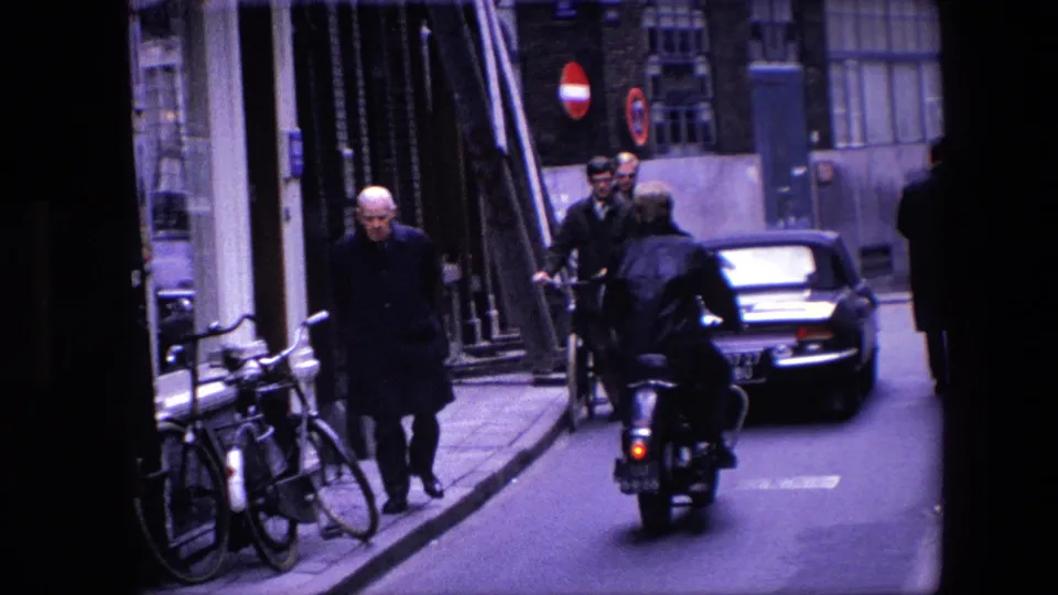 1967 Amsterdam street view showing a motorcycle rider following a dark classic car while pedestrians pass buildings and bicycles line the curb.