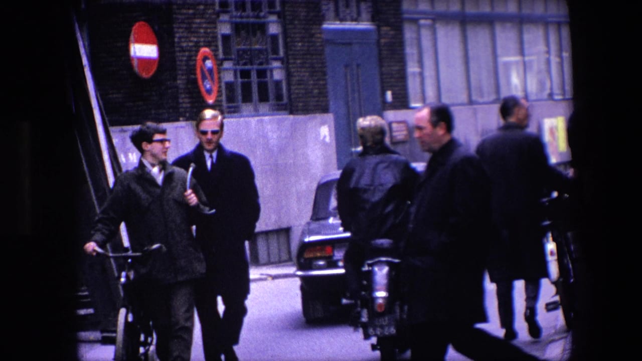 1967 Amsterdam street frame with two men in dark coats near a bicycle, a motorcycle rider moving away, pedestrians, cars, and red no-entry signs in the background.